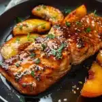 Glazed Bourbon Peach Roasted Salmon fillet with juicy golden peach slices and fresh parsley, shimmering with a caramelized finish, served in a dark skillet.