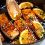 Orange Glazed Salmon fillets in glossy, vibrant orange sauce, garnished with fresh orange slices and green herbs in a skillet, showcasing their juicy texture and colorful presentation.